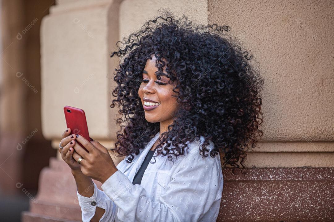 Jovem morena de cabelo encaracolado andando usando o celular. Mensagens de texto na rua. Cidade grande.