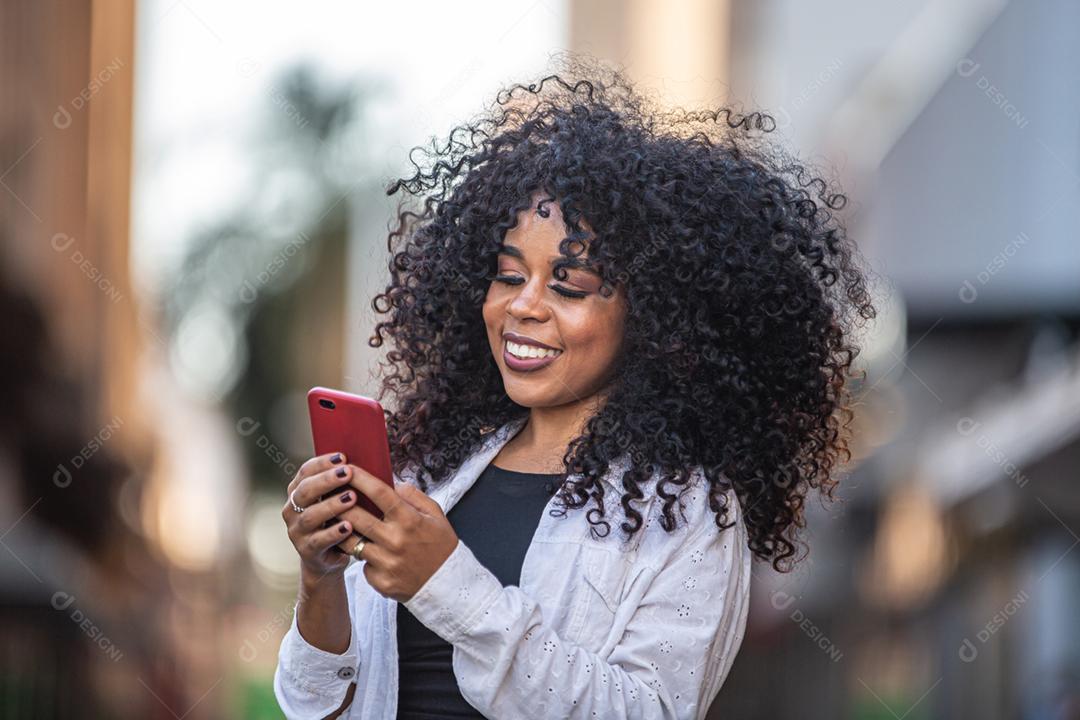 Jovem morena de cabelo encaracolado andando usando o celular. Mensagens de texto na rua. Cidade grande.