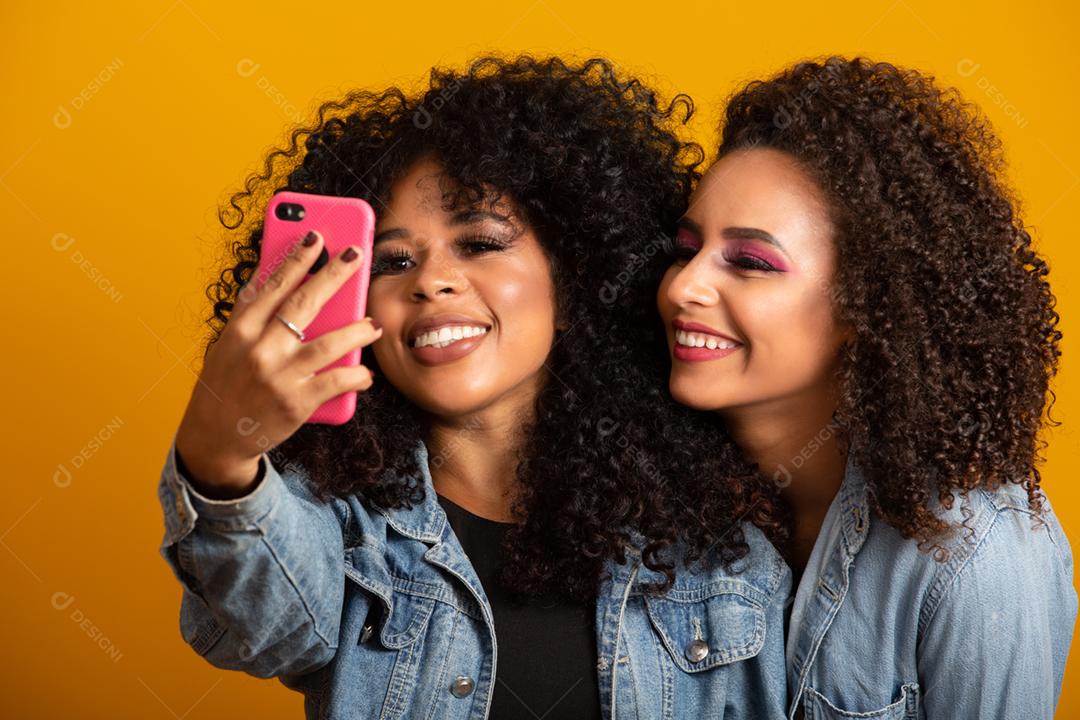 Duas garotas afro-americanas tirando fotos de selfie com seu celular em fundo amarelo