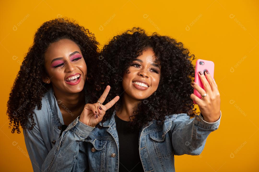Duas garotas afro-americanas tirando fotos de selfie com seu celular em fundo amarelo