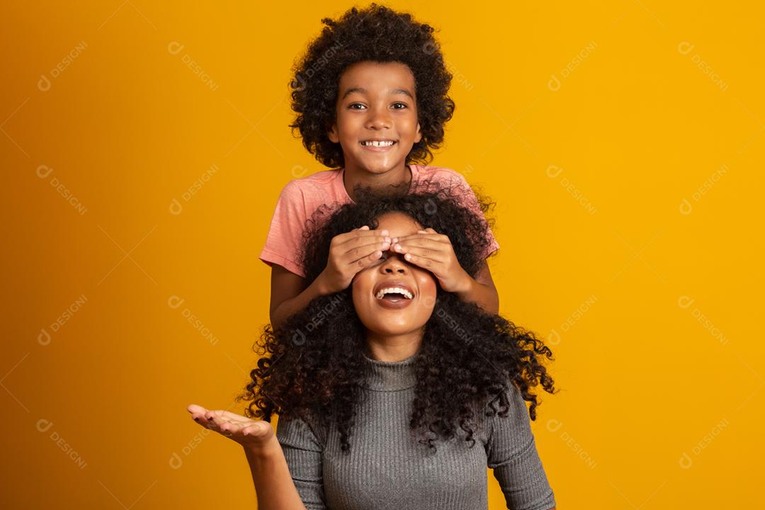 Menino afro-americano segurando os olhos fechados da mãe. Fundo amarelo. Dia das Mães