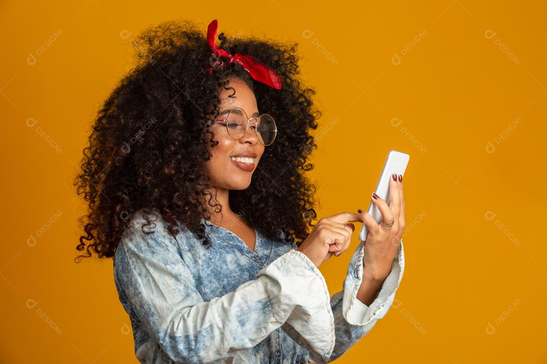 Foto de estúdio de entretida e feliz garota afro-americana com penteado afro segurando smartphone