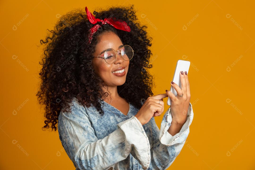 Foto de estúdio de entretida e feliz garota afro-americana com penteado afro segurando smartphone