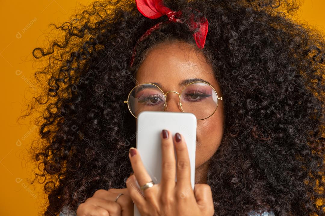 Foto de estúdio de entretida e feliz garota afro-americana com penteado afro segurando smartphone