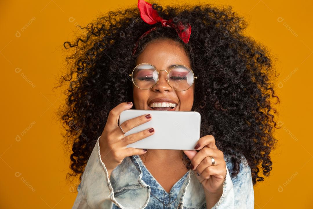 Foto de estúdio de entretida e feliz garota afro-americana com penteado afro segurando smartphone