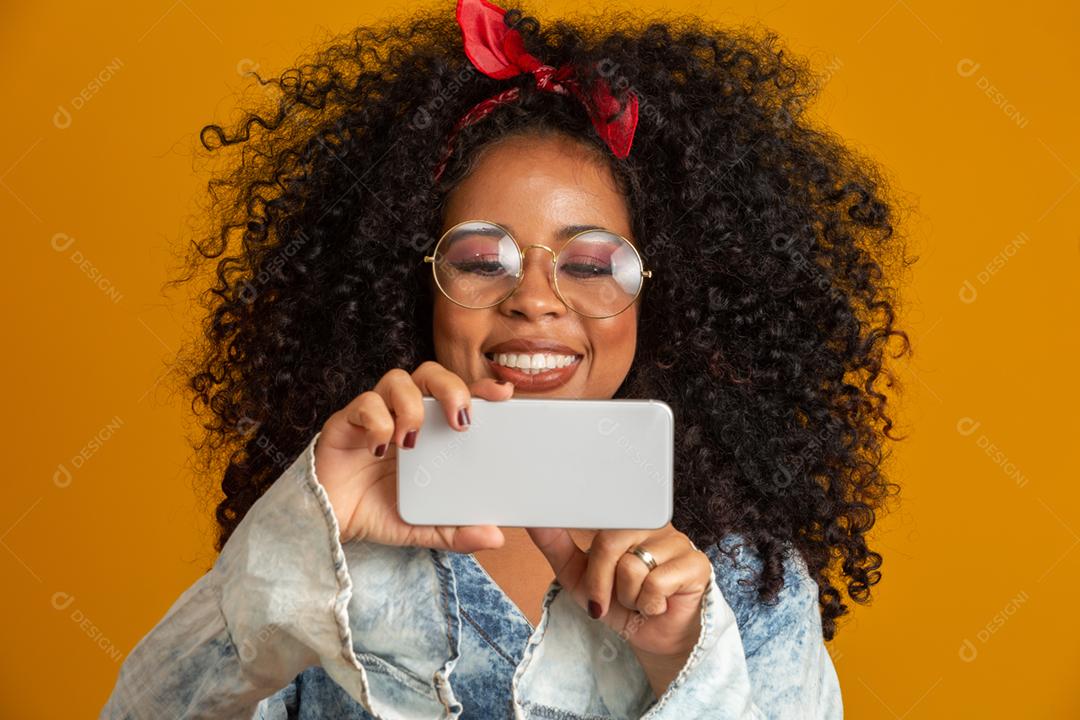 Foto de estúdio de entretida e feliz garota afro-americana com penteado afro segurando smartphone