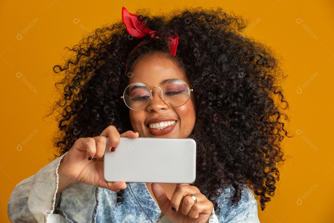 Foto de estúdio de entretida e feliz garota afro-americana com penteado afro segurando smartphone