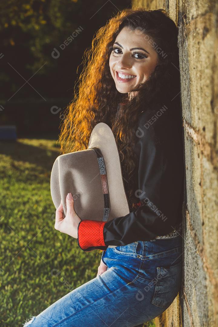 Mulher encostada na parede segurando o seu chapéu