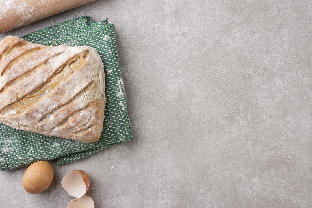 Pão inteiro sobre pano com farinha, ovos e rolo sobre fundo de concreto. Vista do topo.