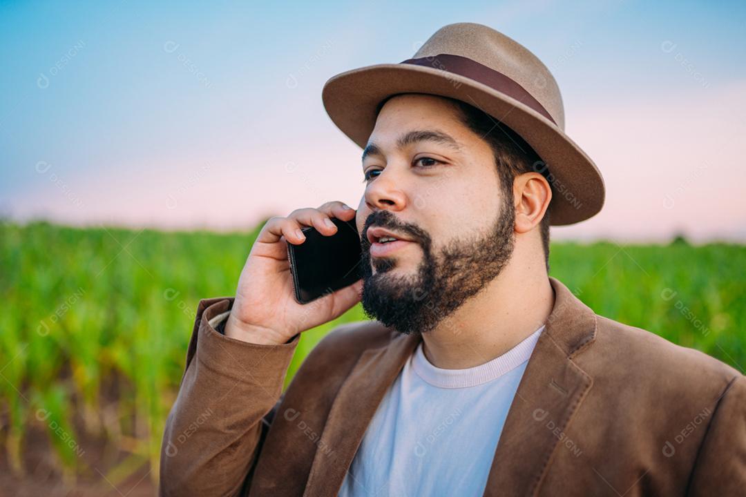 Agricultor latino-americano trabalhando na plantação. Jovem falando no celular, usando chapéu