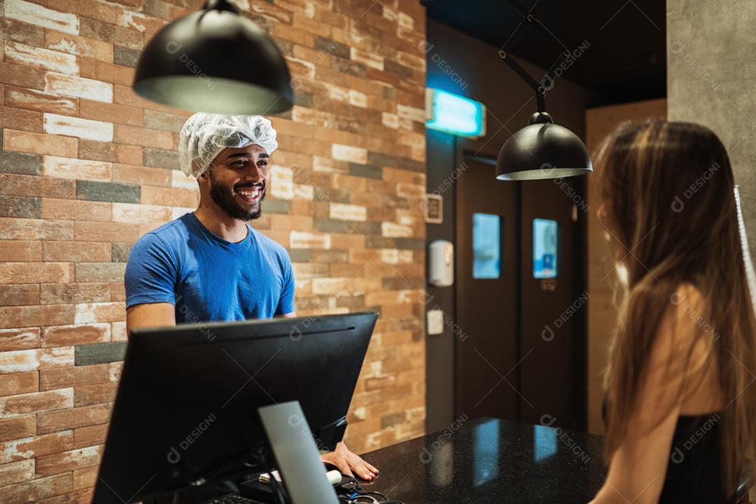 Balconista latina com um boné na cabeça anotando o pedido de uma cliente.