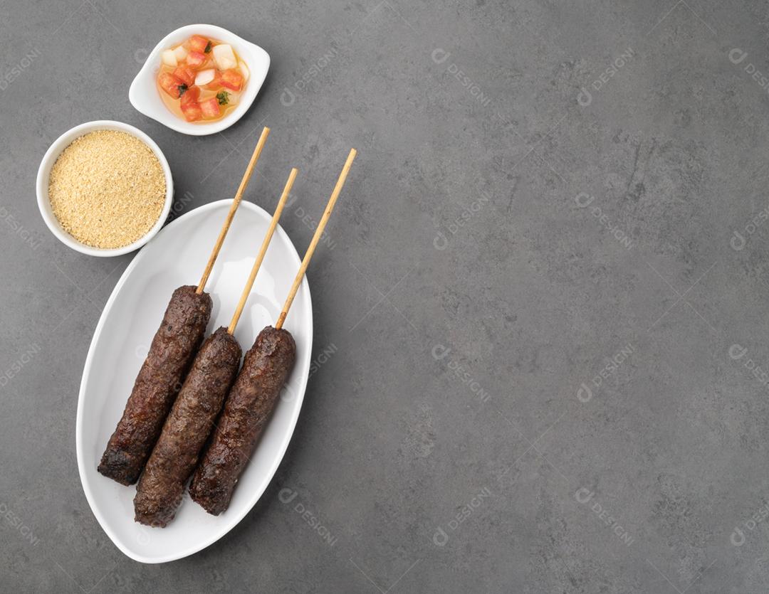 Espetinhos de kafta com farofa, vinagrete sobre fundo de pedra.