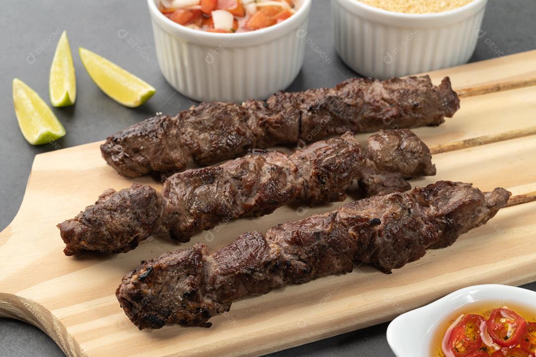 Espetinhos de Kafta de carne sobre tábua de madeira com farofa e vinagrete.