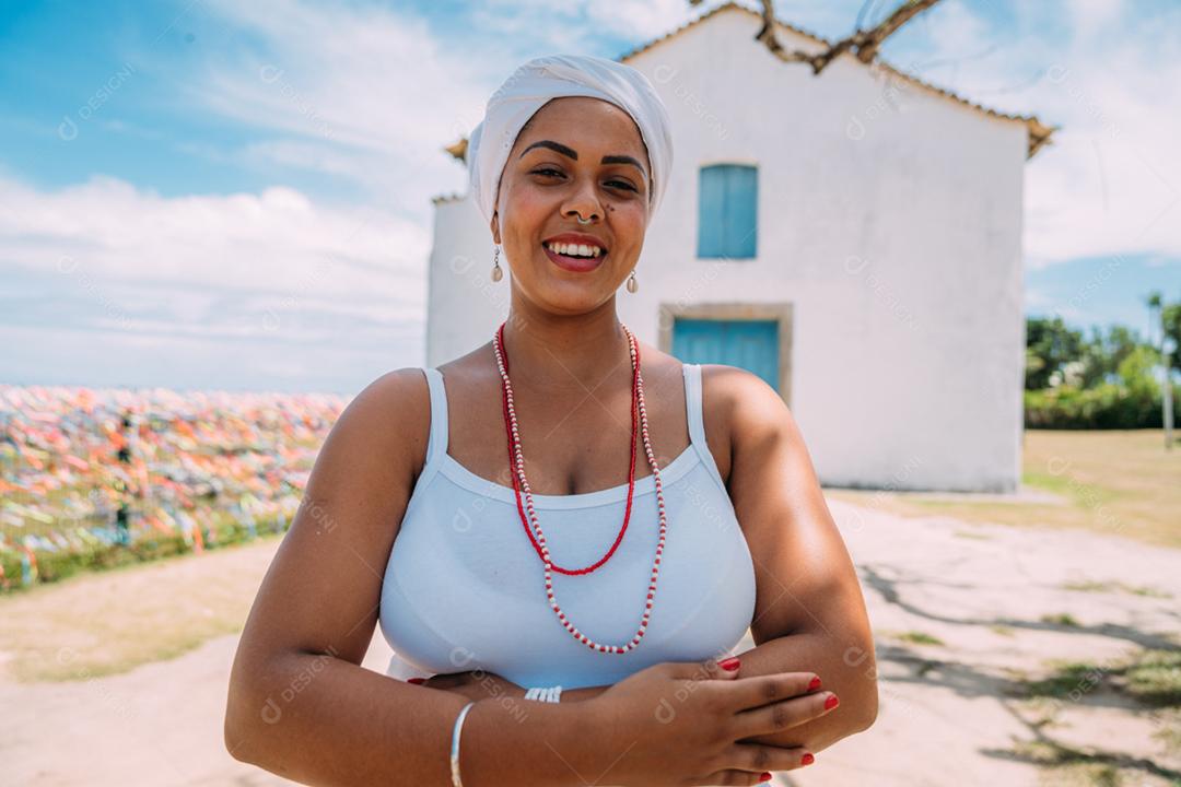 Feliz mulher brasileira vestida com traje tradicional baiano, braços cruzados no centro histórico de Porto Seguro ao fundo