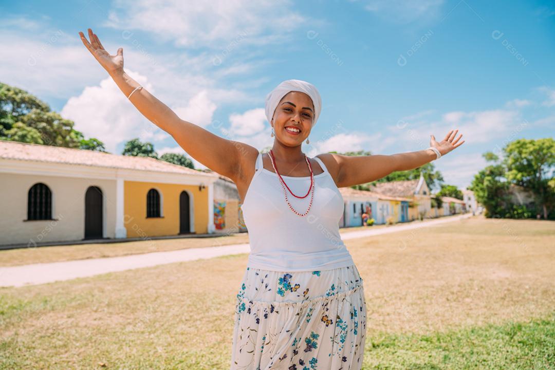 Feliz mulher brasileira vestida com traje tradicional baiano, braços cruzados no centro histórico de Porto Seguro ao fundo