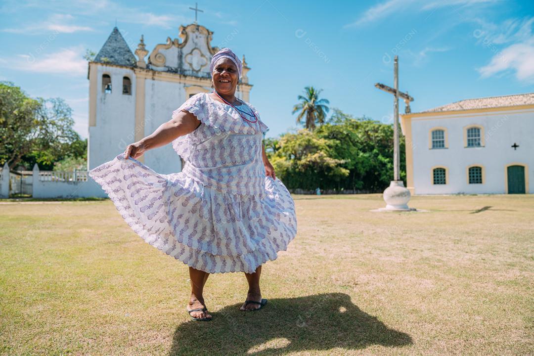 Feliz mulher brasileira de ascendência africana vestida com vestido tradicional baiano dançando em frente à igreja