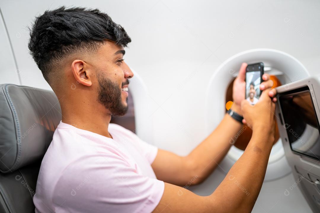 Jovem latino-americano sorrindo fazendo uma chamada de vídeo em seu celular, sentado no avião perto da janela.
