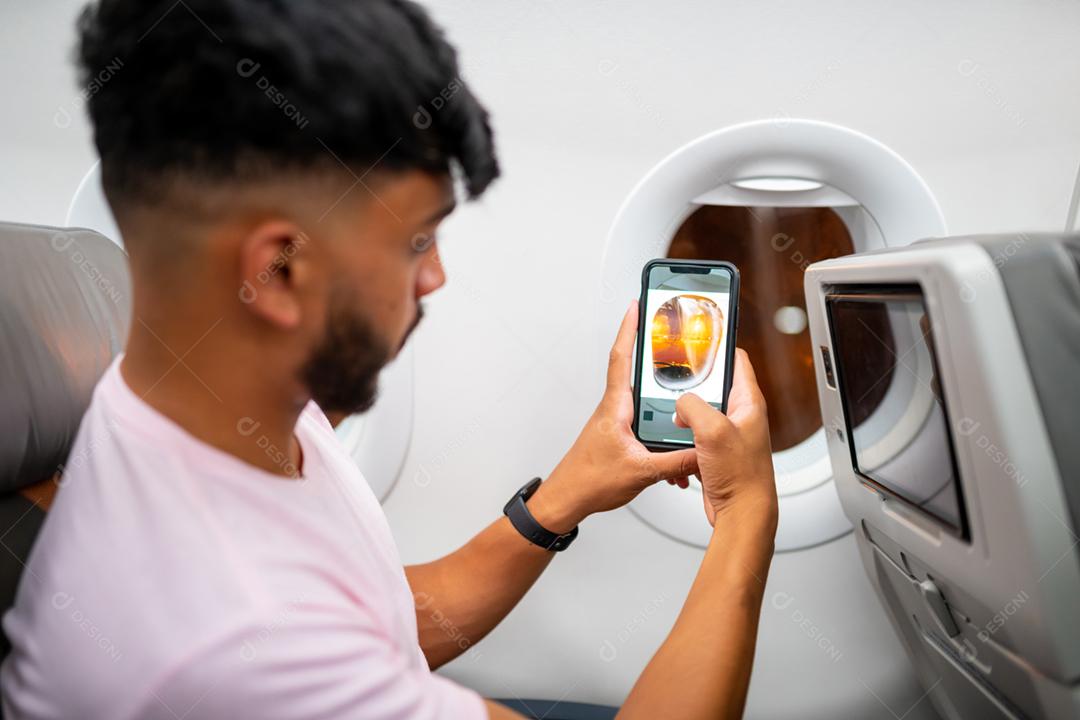 Jovem latino-americano fazendo uma chamada de vídeo no celular, sentado no avião perto da janela.