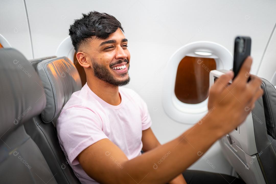 Jovem latino-americano sorrindo fazendo uma chamada de vídeo em seu celular, sentado no avião perto da janela.