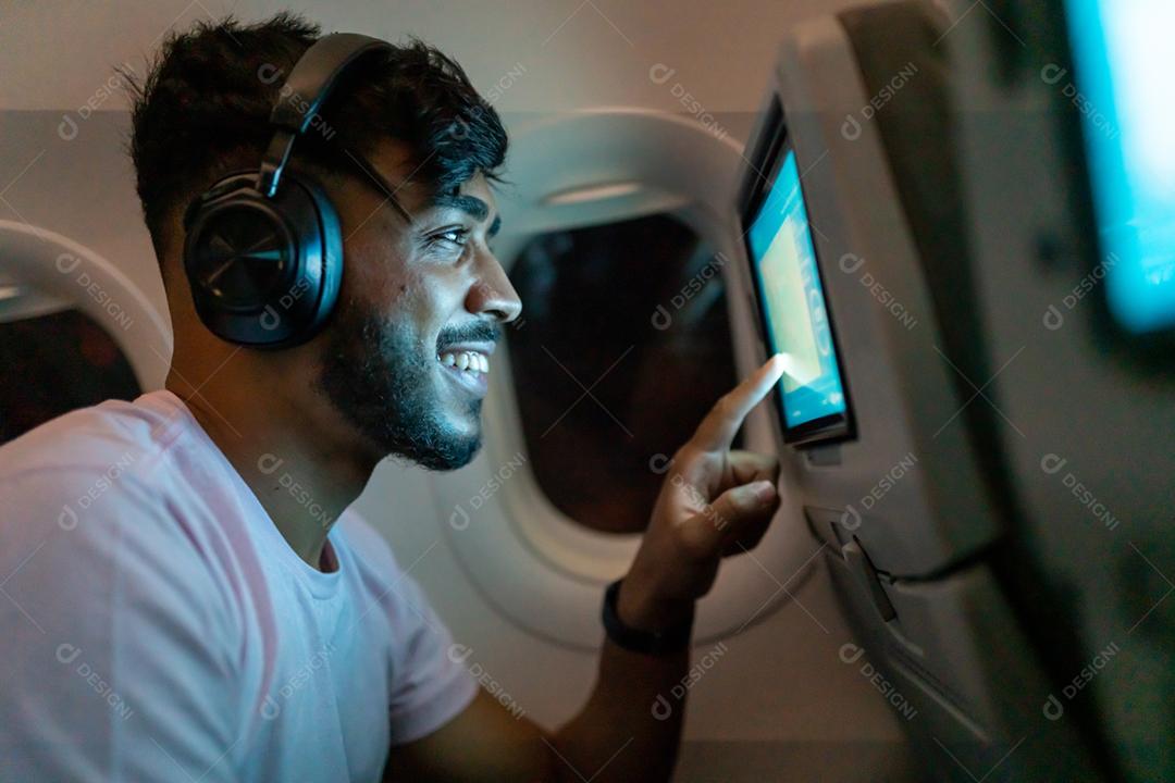 Passageiro no avião tocando a tela de entretenimento LCD. Homem latino-americano na cabine de avião usando dispositivo inteligente ouvindo música em fones de ouvido.