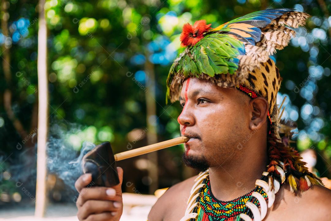Chefe índio da tribo Pataxó fumando cachimbo. Índio brasileiro com cocar de penas e colar
