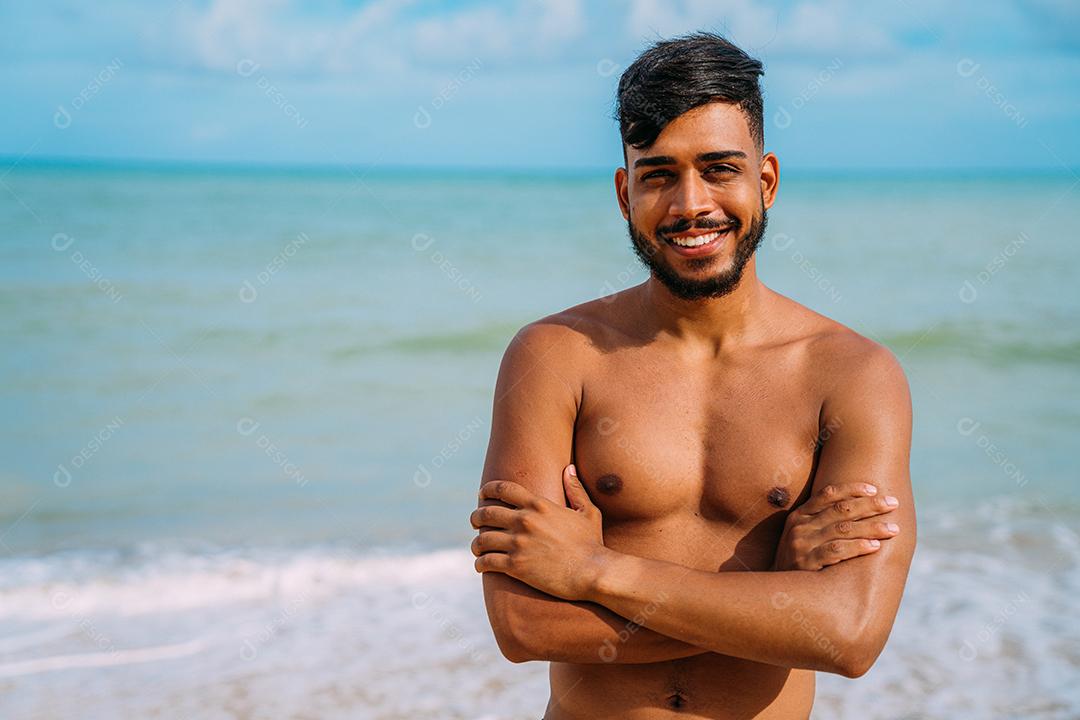 Jovem latino-americano atlético na praia com os braços cruzados