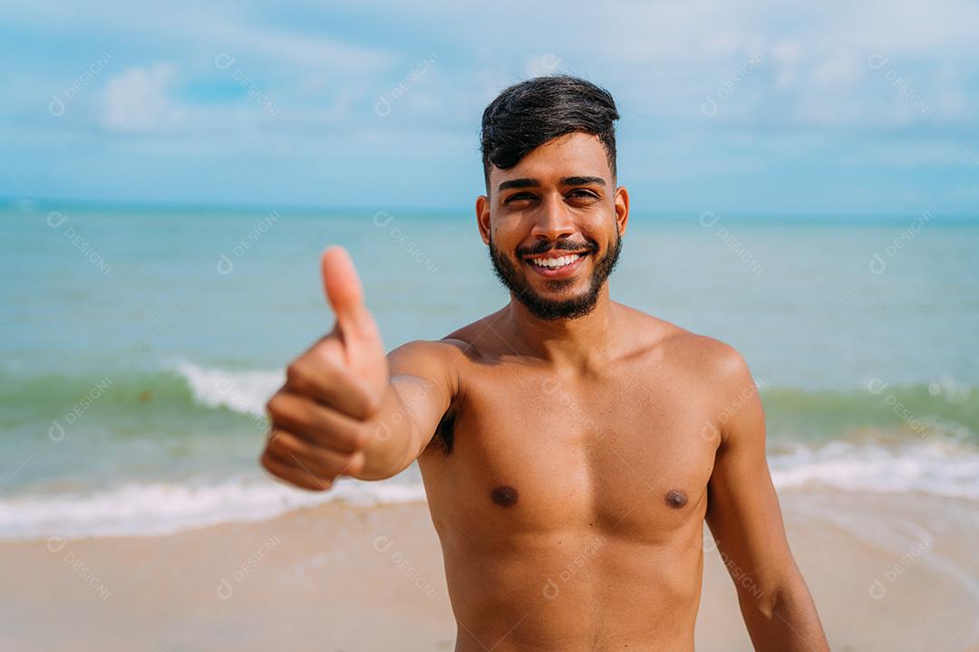 Jovem latino-americano atlético na praia com o polegar para cima