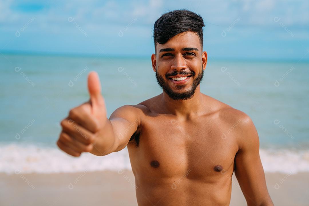 Jovem latino-americano atlético na praia com o polegar para cima