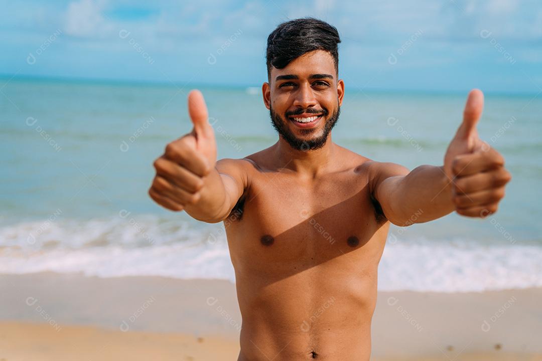 Jovem latino-americano atlético na praia com o polegar para cima