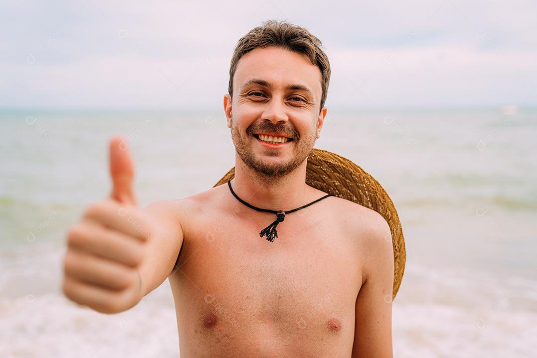 jovem latino-americano na praia com um chapéu e polegar