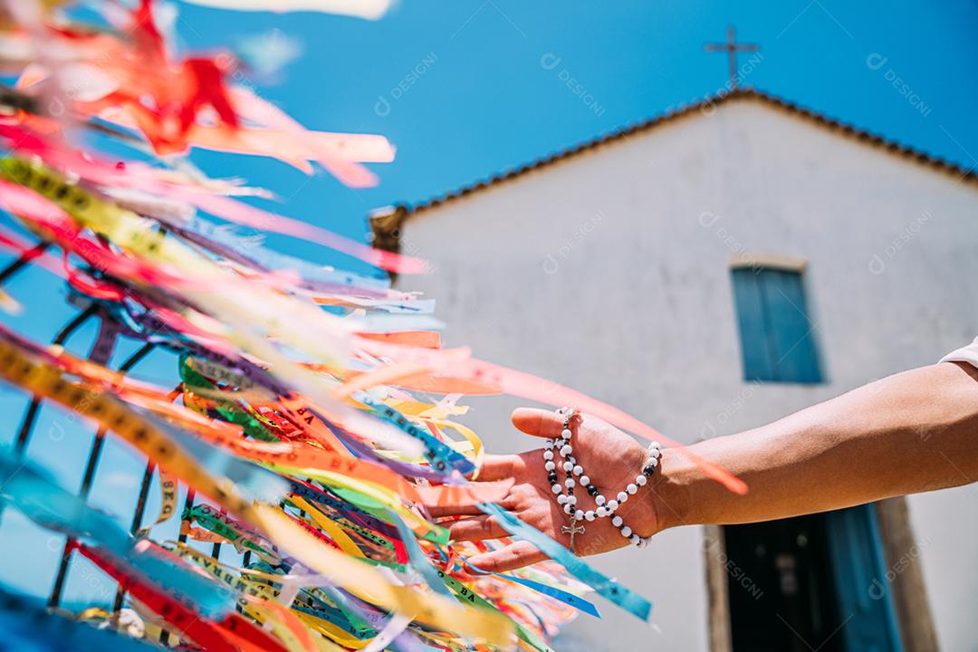 A mão de um homem fazendo um pedido com fitas brasileiras na cerca de uma igreja em Arraial dAjuda