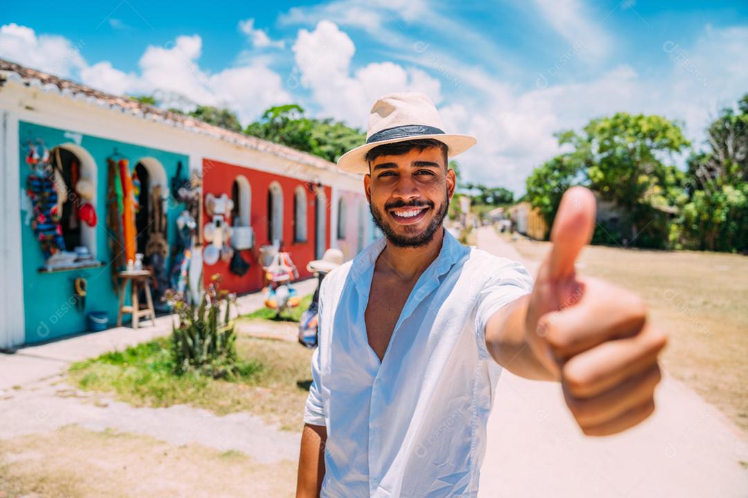 Turista fazendo selfie no centro histórico de Porto Seguro. Homem latino-americano de chapéu e polegar para cima