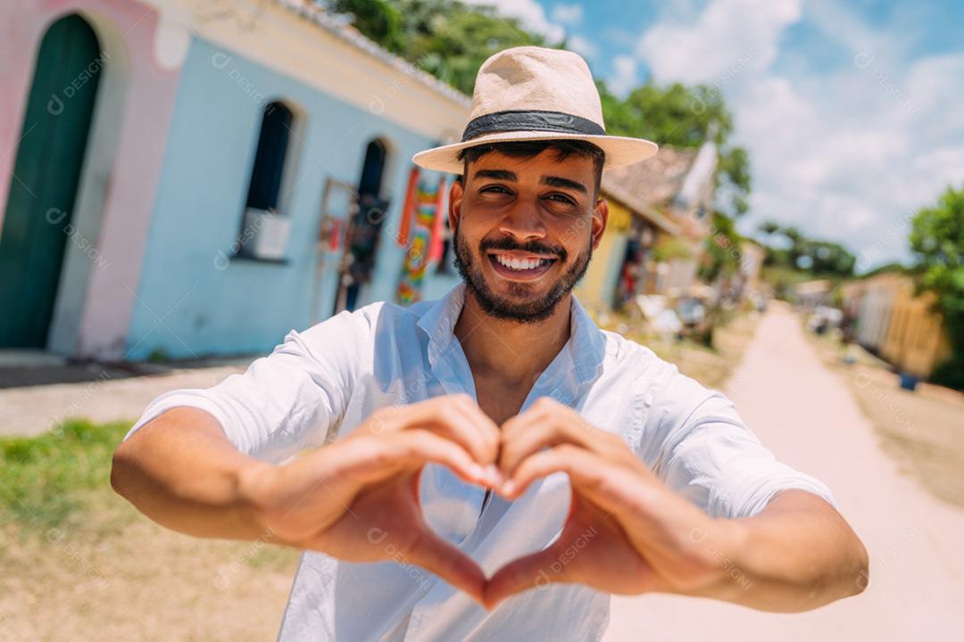 Homem latino-americano de chapéu, fazendo um coração com as mãos, sorrindo para a câmera
