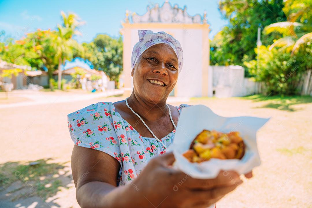 Mulher oferecendo acarajé - comida típica baiana - no centro histórico de Porto Seguro ao fundo