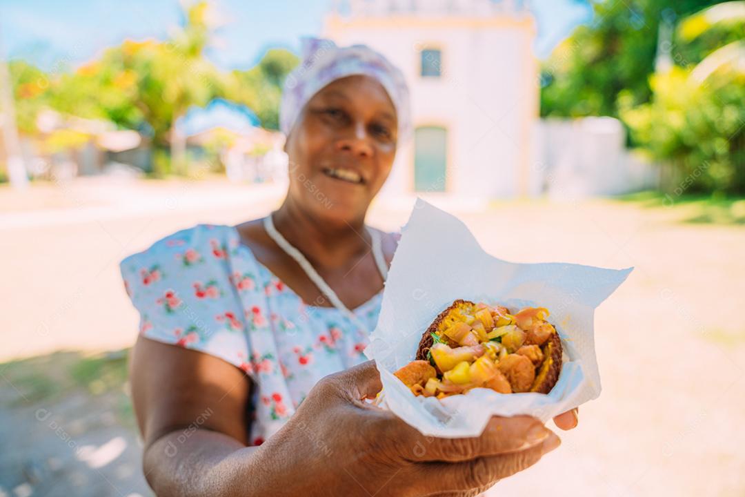 Mulher oferecendo acarajé - comida típica baiana - no centro histórico de Porto Seguro ao fundo