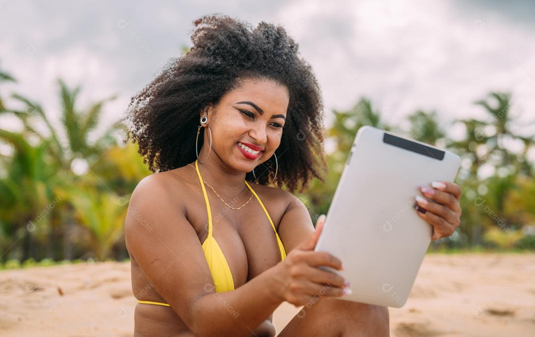 Férias de verão, tecnologia e conceito de internet. mulher latino-americana sentada na areia, tomando sol na praia com um tablet