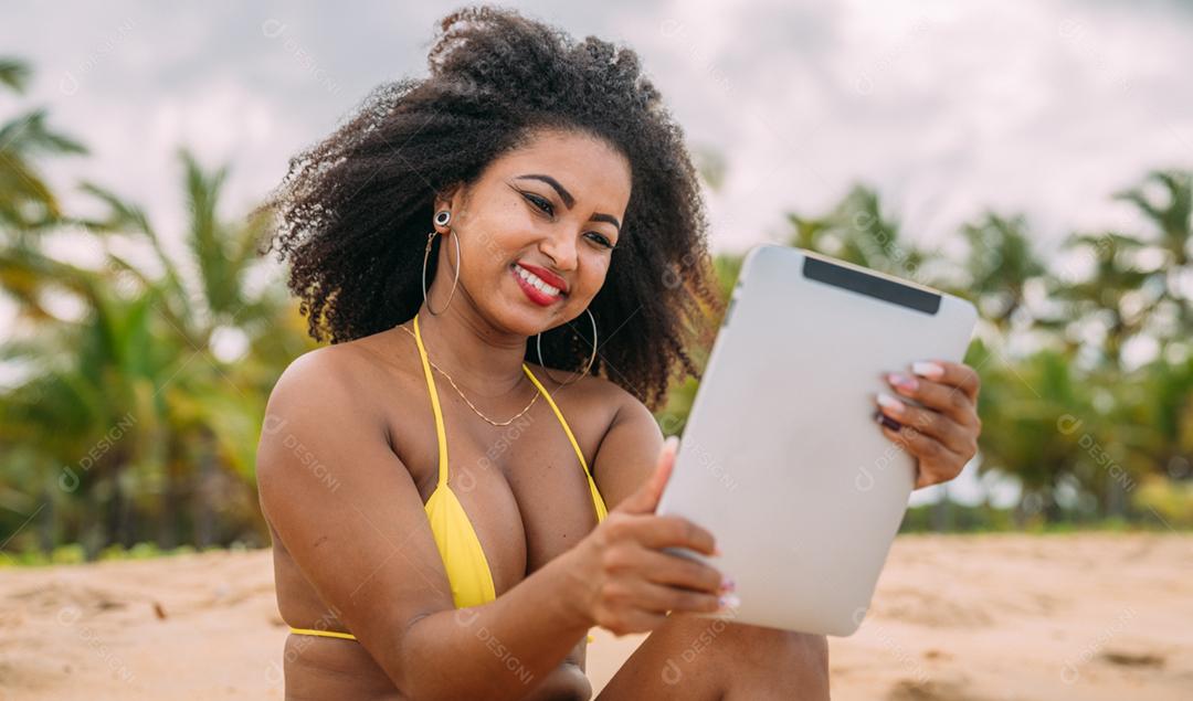 Férias de verão, tecnologia e conceito de internet. mulher latino-americana sentada na areia, tomando sol na praia com um tablet