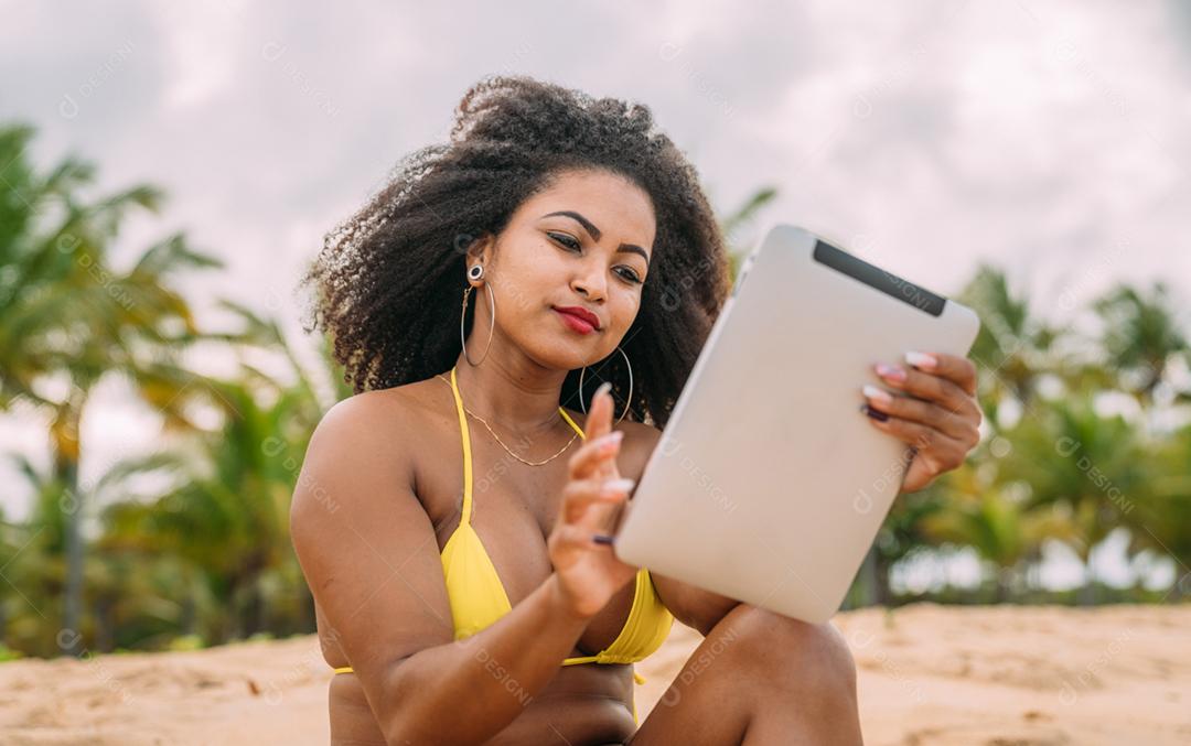 Férias de verão, tecnologia e conceito de internet. mulher latino-americana sentada na areia, tomando sol na praia com um tablet