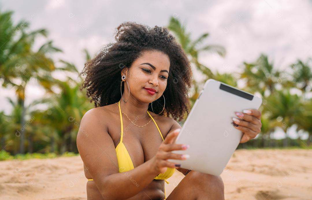 Férias de verão, tecnologia e conceito de internet. mulher latino-americana sentada na areia, tomando sol na praia com um tablet