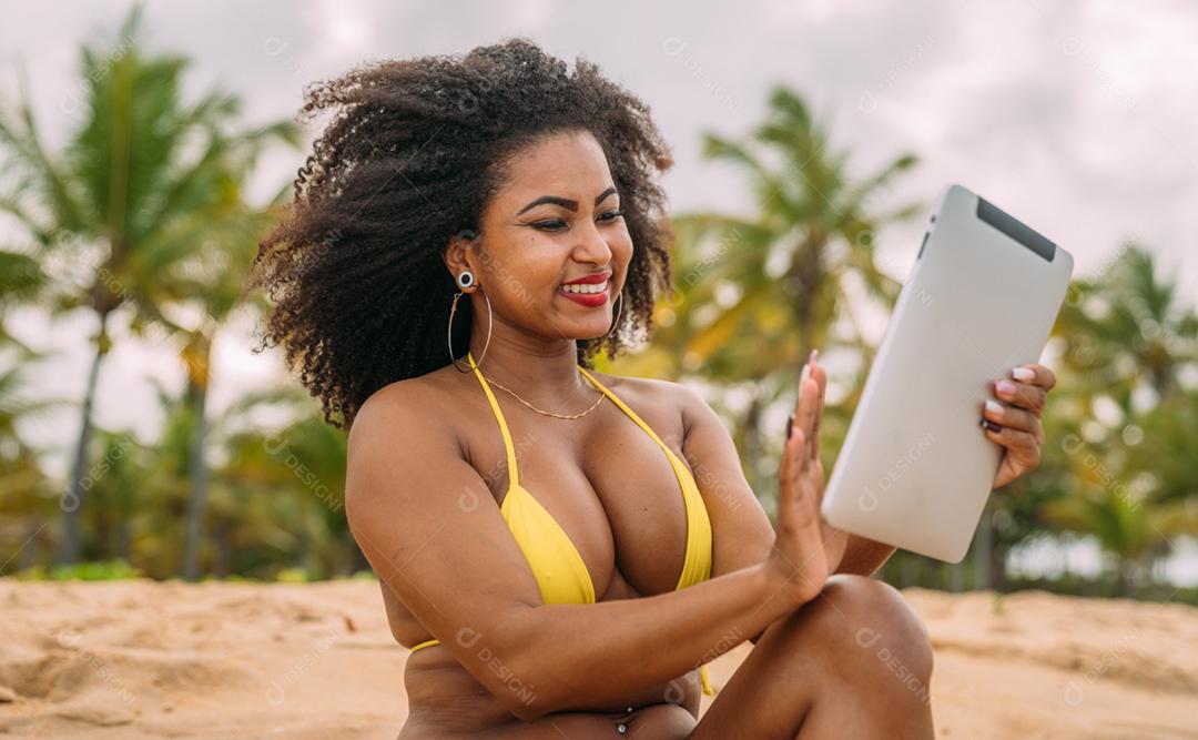 Férias de verão, tecnologia e conceito de internet. mulher latino-americana sentada na areia, tomando sol na praia com um tablet