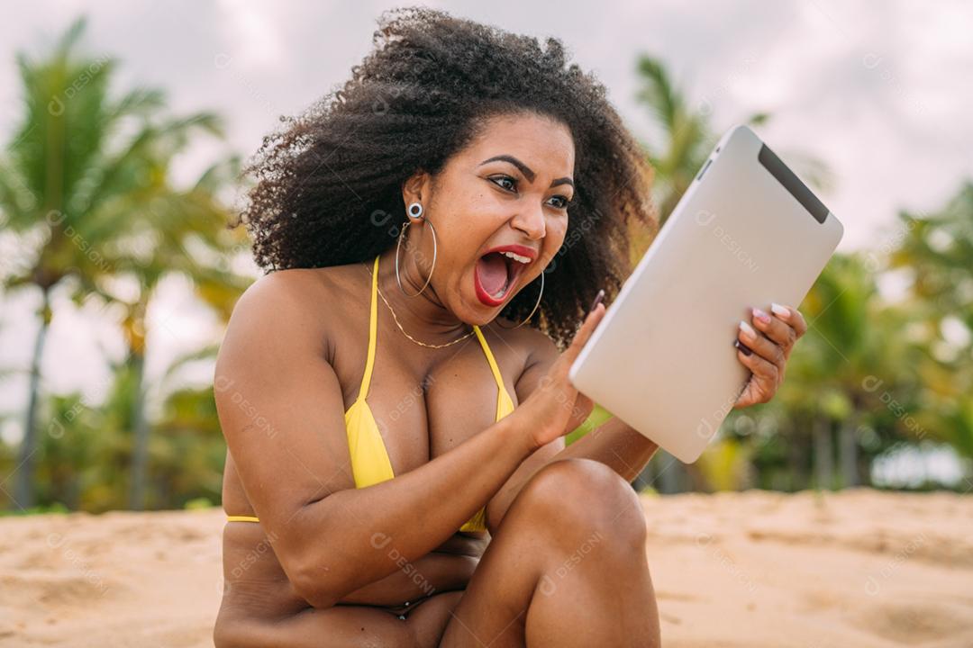 Summer vacation, technology and internet concept. Latin American woman sitting on the sand, sunbathing on the beach with a tablet