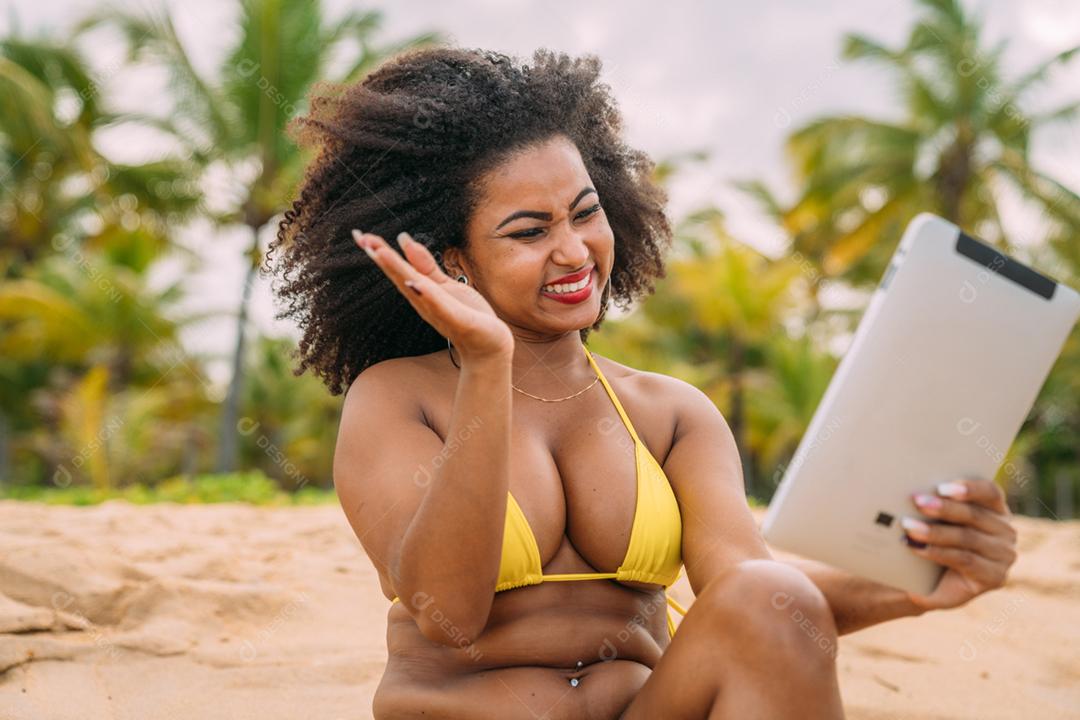 Férias de verão, tecnologia e conceito de internet. mulher latino-americana sentada na areia, tomando sol na praia com um tablet