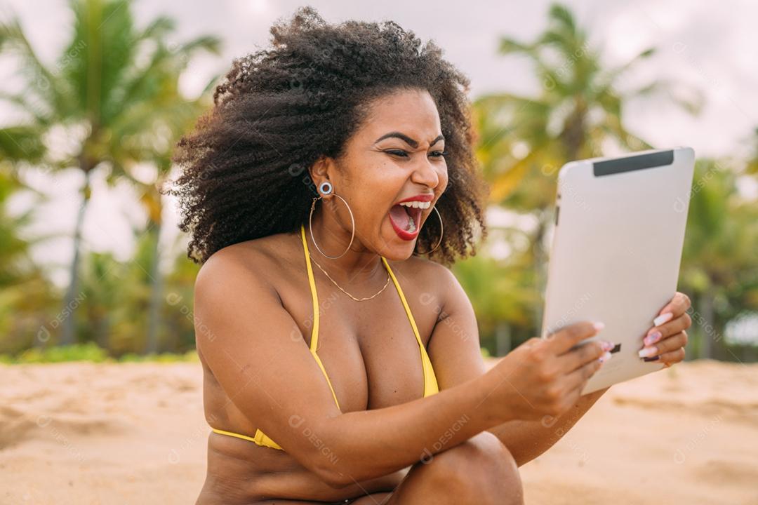 Férias de verão, tecnologia e conceito de internet. mulher latino-americana sentada na areia, tomando sol na praia com um tablet