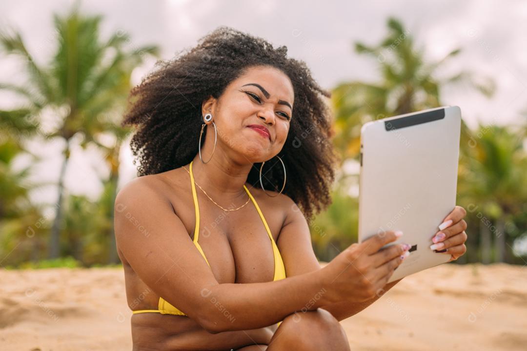 Férias de verão, tecnologia e conceito de internet. mulher latino-americana sentada na areia, tomando sol na praia com um tablet