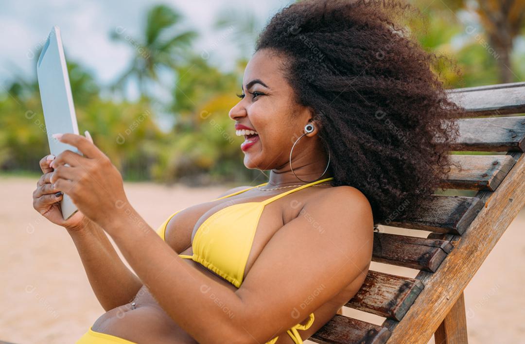 Mulher fazendo uma chamada de vídeo nas férias de verão. mulher latino-americana sentada na cadeira de praia com um tablet