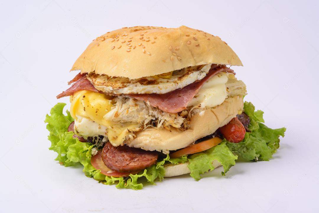 Sanduíche de hambúrguer grande com presunto defumado, queijo, ovos, salsicha, bacon, alface e tomate em fundo branco.