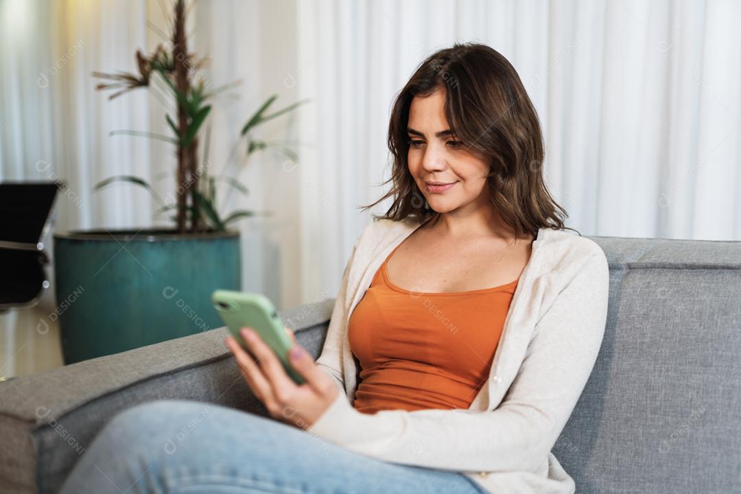 Feliz dama latina relaxa em casa sozinha, sente-se na sala e compartilhe boas notícias nas mídias sociais via celular