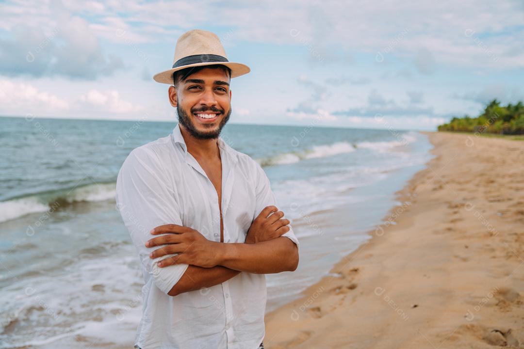 Amigável jovem latino-americano com os braços cruzados na praia, usando um chapéu