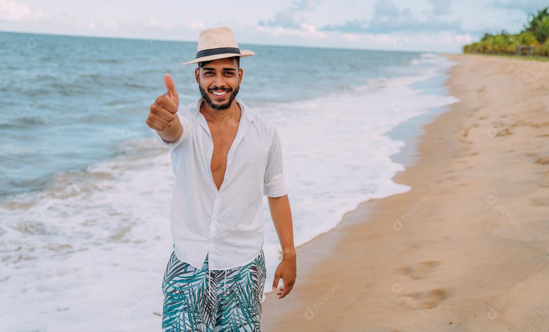 Amigável jovem latino-americano fazendo gesto de polegar para cima na praia, usando um chapéu