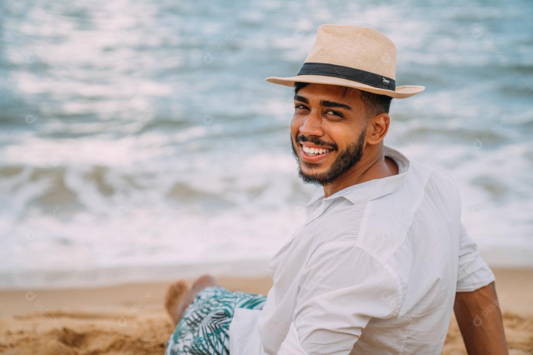 Silhueta de um jovem na praia. homem latino-americano sentado na areia da praia, usando um chapéu o